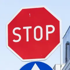 Znak b20 STOP. Czerwony ośmiokąt z białym napisem STOP. W tle błękitne niebo i fragment budynku.