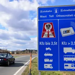 Niebieski znak drogowy z informacją o winietach i opłatach drogowych w Austrii, obok drogi z samochodem.