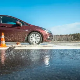 Czerwony samochód na mokrej nawierzchni, obok pomarańczowy pachołek. Woda rozpryskuje się spod kół. To pokazuje, jak działa asr co to za system.