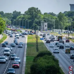 Tłoczna trasa w Warszawie, pełna samochodów i autobusów, z zielenią po bokach.