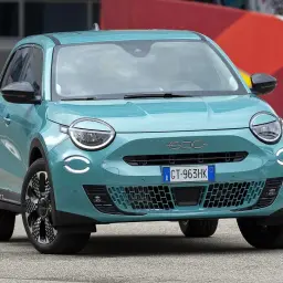 Nowy, turkusowy Fiat 600 z okrągłymi światłami i charakterystycznym grillem, zaparkowany na tle czerwonej ściany z graffiti.