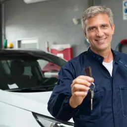 Uśmiechnięty mechanik w granatowym kombinezonie wręcza kluczyki do samochodu, symbolizując pomyślną homologację i odbiór pojazdu.