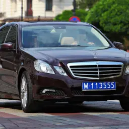 Ciemnobrązowy Mercedes z tablicą W 113555, wskazującą na rejestracje dyplomatyczne, jedzie po ulicy.
