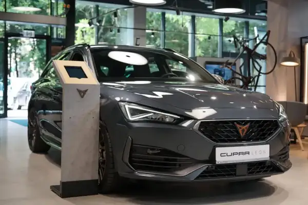Nowoczesne konie mechaniczne, szary Cupra Leon, prezentują się w salonie. Obok stoi multimedialny kiosk.