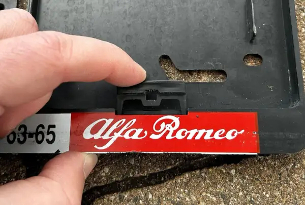 Palec naciska na zatrzask, pokazując, jak zdjąć tablice rejestracyjne z ramki Alfa Romeo.