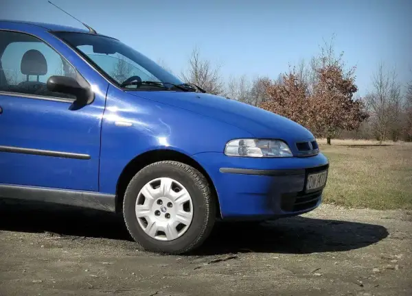 Błękitny Fiat Albea stoi na polnej drodze. W tle widać drzewa i błękitne niebo.
