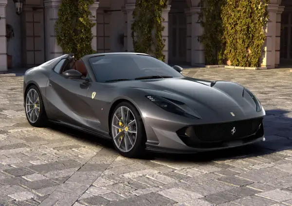 Szary kabriolet Ferrari 812 GTS z żółtymi zaciskami hamulcowymi stoi na brukowanym dziedzińcu przed budynkiem porośniętym bluszczem.