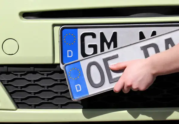 Dłoń trzyma tablice rejestracyjne państw. Jedna z nich ma oznaczenie "GM", druga "OE".